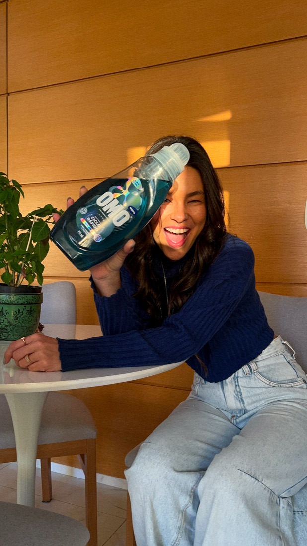Carol rindo em casa, sentada à mesa ao lado de uma planta
